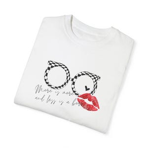 Puede incluir: Camiseta blanca con un gráfico de gafas de cuadros blanco y negro y una impresión de labios rojos. El texto "More is more and less is a bore" está impreso en la camiseta.