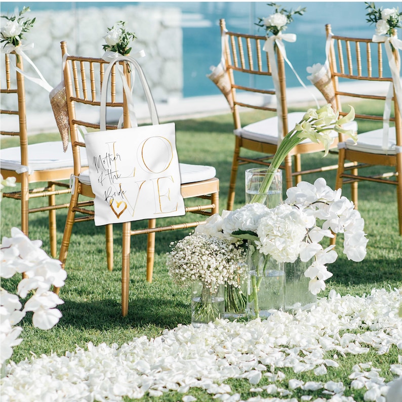 Puede incluir: Bolsa de tela blanca con letras doradas que dicen "Mother of the Bride LOVE" con un coraz&oacute;n dorado en el centro. La bolsa est&aacute; sentada en una silla dorada con flores blancas y p&eacute;talos en el suelo.