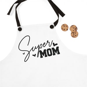 Puede incluir: Delantal blanco con tirantes ajustables negros y botones negros. El delantal tiene el texto "Super Mom" en negro con corazones.