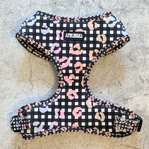 Può includere: Imbracatura per cani con motivo a quadri bianchi e neri e un design con stampa leopardata in rosa, pesca e lavanda. L'imbracatura riporta la scritta "PUP ROCK PET ACCESSORIES" su un'etichetta nera.