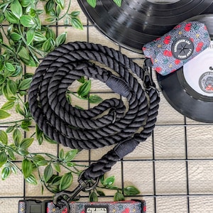 May include: A black rope dog lead and matching collar set with a cherry and lightning bolt pattern. The set includes a black lead with a metal clasp and a collar with a plastic buckle. A matching waste bag dispenser is also included.