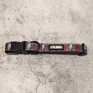 Può includere: Un collare per cani nero con sfondo marrone e motivo floreale. Il collare ha una fibbia nera e un anello argentato. Il testo "Pup Rock Pet Accessories" è stampato sul collare.