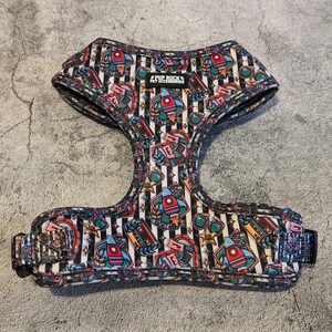May include: A black and white striped dog harness featuring a colourful rocket ship, cassette tape, and other retro themed designs. The harness has the text "Pup Rock Pet Accessories" on it.