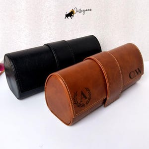 Könnte beinhalten: Zwei zylindrische Uhrenetuis, eines in Schwarz und eines in Braun, werden gezeigt. Das braune Etui hat die Initialen "CW" und "A" mit einem Lorbeerkranz-Design. Beide Etuis haben einen Riemenverschluss.