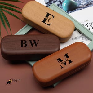 May include: Three wooden eyeglass cases in shades of brown. Each case is rectangular with a curved top and features a personalized initial and name in black. The cases are arranged on a decorative surface.