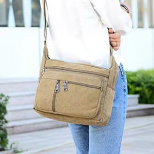 May include: A tan canvas messenger bag with two zippered compartments on the front. The bag has a long adjustable strap and is worn over the shoulder. The bag is a neutral color and appears to be made of a durable material.