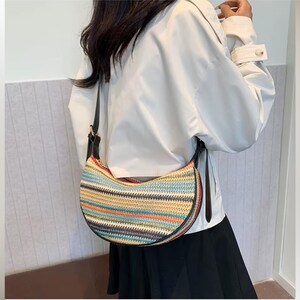 May include: A half-moon shaped handbag with a woven design in various colors, including blue, orange, and green. The bag has a black strap and trim. The bag is worn over the shoulder.