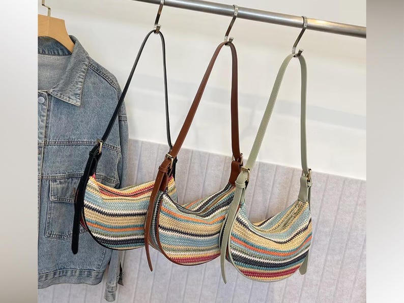 May include: Three shoulder bags with striped patterns in various colors, including blue, red, and yellow, hang from a metal bar. Each bag has a different strap color: black, brown, and light green. A denim jacket hangs on a wooden hanger to the left.