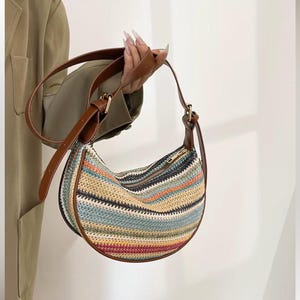 May include: A half-moon shaped handbag with a woven, striped pattern in various colors, including blue, orange, and red. The bag has a brown leather strap and trim, and a zipper closure. The bag is held by a person.