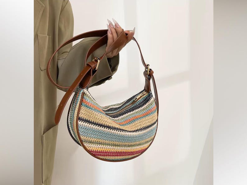May include: A half-moon shaped handbag with a woven, striped pattern in various colors, including blue, orange, and red. The bag has a brown leather strap and trim, and a zipper closure. The bag is held by a person.