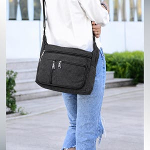 May include: A dark gray canvas crossbody bag featuring a rectangular shape, an adjustable shoulder strap, and two front zippered pockets with silver-toned metal hardware. The bag has a textured fabric finish and a main top zipper closure.