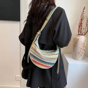 May include: A colorful, woven crescent-shaped handbag with a light green strap. The bag features horizontal stripes in various colors, including blue, red, yellow, and orange. The strap is adjustable and appears to be made of a smooth material.