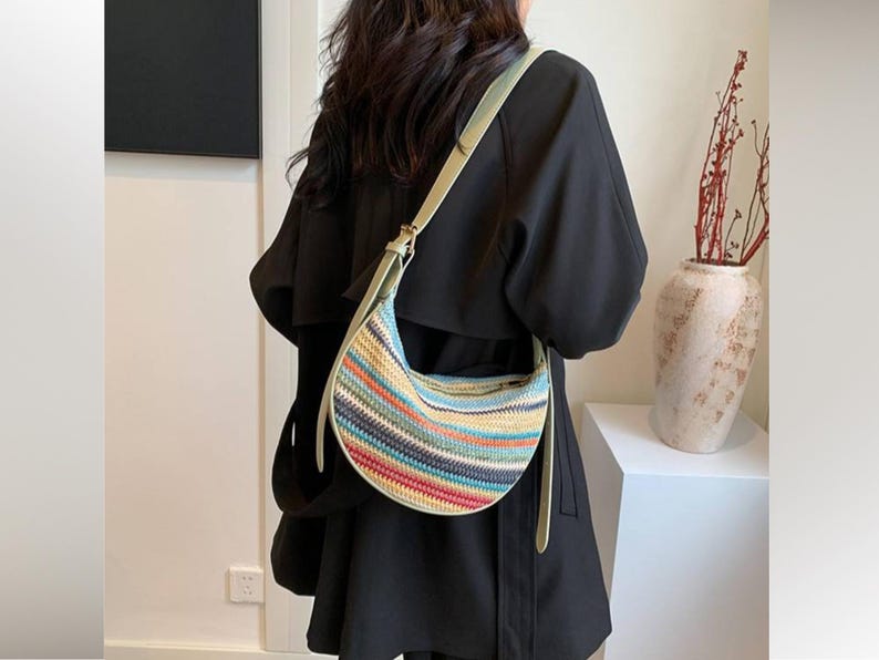 May include: A colorful, woven crescent-shaped handbag with a light green strap. The bag features horizontal stripes in various colors, including blue, red, yellow, and orange. The strap is adjustable and appears to be made of a smooth material.
