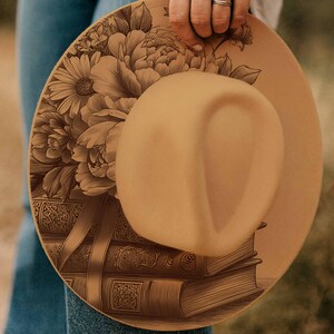 K&ouml;nnte beinhalten: Ein beigefarbener Fedora-Hut mit einem detaillierten Blumen- und Buchdesign. Der Hut hat eine breite Krempe und eine abgerundete Krone. Das florale Design umfasst Pfingstrosen und G&auml;nsebl&uuml;mchen. Die B&uuml;cher sind mit dekorativem Band gestapelt.