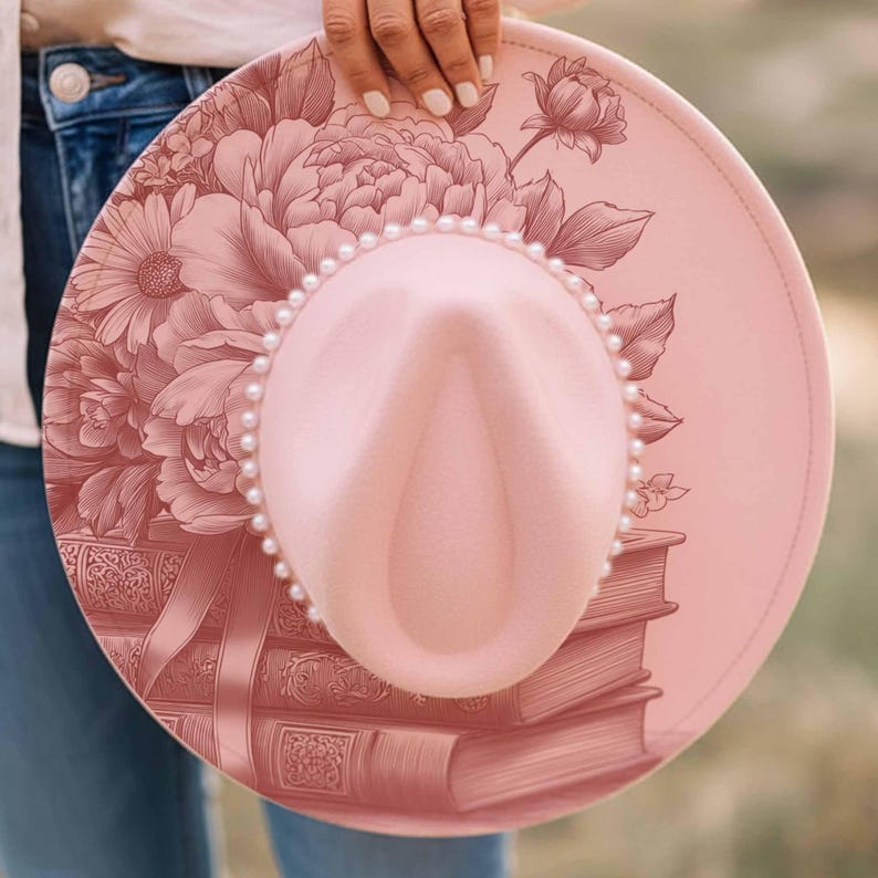 K&ouml;nnte beinhalten: Ein rosa Fedora-Hut mit floralem Design und Perlenbesatz. Der Hut zeigt einen Stapel B&uuml;cher und ein gro&szlig;es Blumenmuster in einem dunkleren Rosaton. Der Hut wird von einer Person gehalten.