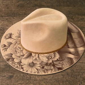 K&ouml;nnte beinhalten: Ein cremefarbener Hut im Fedora-Stil mit Blumen- und Buchdesign. Der Hut hat ein hellbraunes Seilband und eine breite Krempe. Die Krempe ist mit einem detaillierten Blumenmuster auf der linken Seite und einem Buchdesign auf der rechten Seite verziert.
