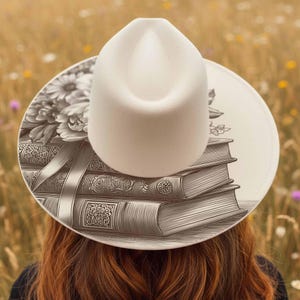 K&ouml;nnte beinhalten: Ein cremefarbener Fedora-Hut mit breiter Krempe, mit einer detaillierten Schwarz-Wei&szlig;-Illustration von B&uuml;chern und Blumen. Der Hut befindet sich vor einem verschwommenen Hintergrund eines Feldes.