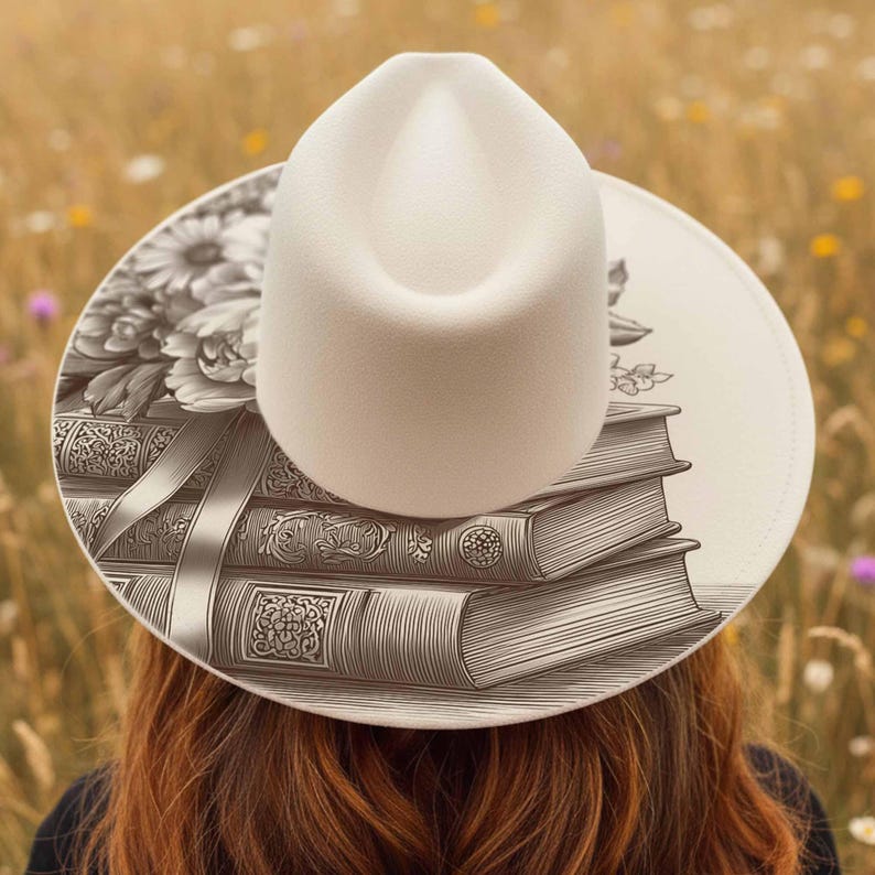 K&ouml;nnte beinhalten: Ein cremefarbener Fedora-Hut mit breiter Krempe, mit einer detaillierten Schwarz-Wei&szlig;-Illustration von B&uuml;chern und Blumen. Der Hut befindet sich vor einem verschwommenen Hintergrund eines Feldes.