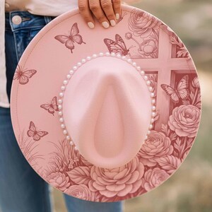 May include: A pink fedora-style hat with a pearl-lined brim. The hat features a cross design, butterflies, and floral patterns. The hat is held by a person.
