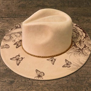 May include: A cream-colored fedora hat with a wide brim featuring a brown floral and butterfly design. The hat has a braided band and is set against a wooden surface.