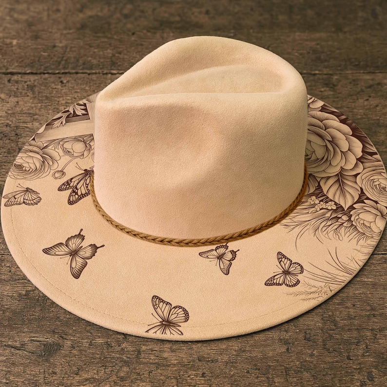 May include: A cream-colored fedora hat with a wide brim featuring a brown floral and butterfly design. The hat has a braided band and is set against a wooden surface.