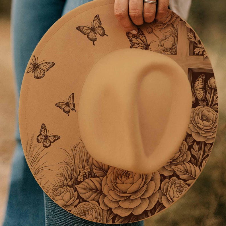 May include: Tan fedora hat with a wide brim, featuring a detailed butterfly and floral design. The hat has a classic teardrop crown and is held by a person wearing a ring. The brim is decorated with butterflies and flowers.