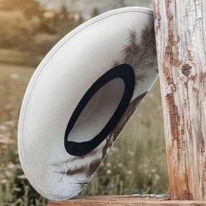 K&ouml;nnte beinhalten: Ein cremefarbener Cowboyhut mit schwarzem Hutband und einem dekorativen Feder- und Baumdesign. Der Hut liegt auf einer Holzoberfl&auml;che, mit einem verwitterten Holzpfosten im Hintergrund. Der Hutrand ist leicht gebogen.