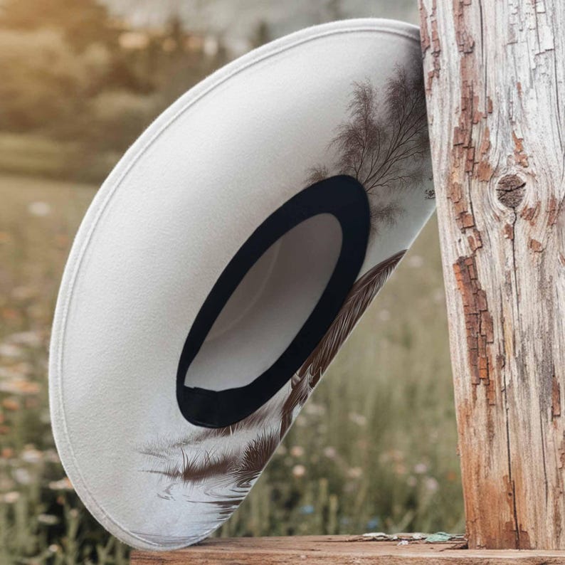 K&ouml;nnte beinhalten: Ein cremefarbener Cowboyhut mit schwarzem Hutband und einem dekorativen Feder- und Baumdesign. Der Hut liegt auf einer Holzoberfl&auml;che, mit einem verwitterten Holzpfosten im Hintergrund. Der Hutrand ist leicht gebogen.