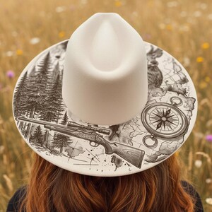May include: A cream-coloured fedora hat featuring a detailed black and white illustration on the brim. The design includes a rifle, compass, map, and forest scene. The hat is set against a blurred background of wildflowers.