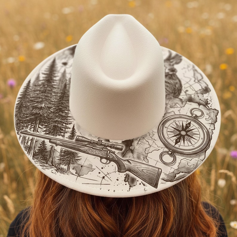 May include: A cream-coloured fedora hat featuring a detailed black and white illustration on the brim. The design includes a rifle, compass, map, and forest scene. The hat is set against a blurred background of wildflowers.