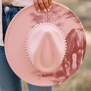 K&ouml;nnte beinhalten: Ein rosa Cowboyhut mit Perlenbesatz und einer detaillierten Illustration eines Pferdes, das Wasser trinkt. Der Hut hat ein Baumdesign. Der Hut wird von einer Person gehalten, die eine blaue Jeans tr&auml;gt.
