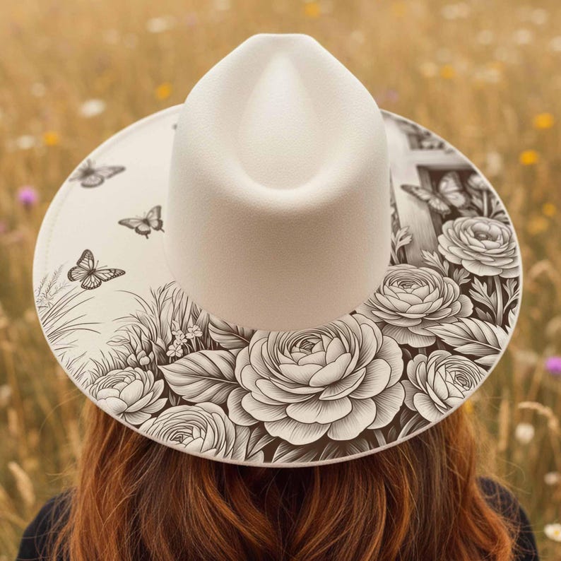 May include: A cream-colored fedora hat with a wide brim featuring a black and white floral and butterfly design. The hat's crown is smooth, and the brim is decorated with detailed illustrations of roses, butterflies, and foliage.