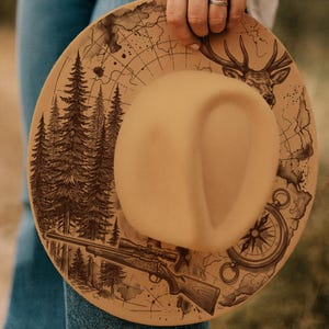 May include: A tan hat with a detailed design featuring a forest, deer, map, compass, and rifle. The hat has a wide brim and a central indentation. The illustrations are in dark brown, giving a rustic, outdoorsy feel.
