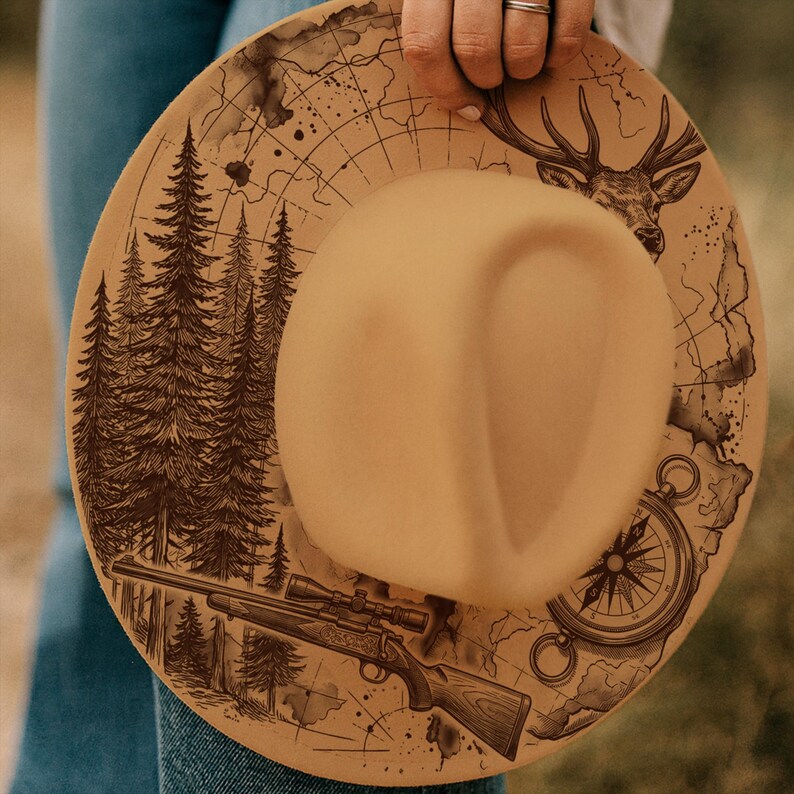 May include: A tan hat with a detailed design featuring a forest, deer, map, compass, and rifle. The hat has a wide brim and a central indentation. The illustrations are in dark brown, giving a rustic, outdoorsy feel.