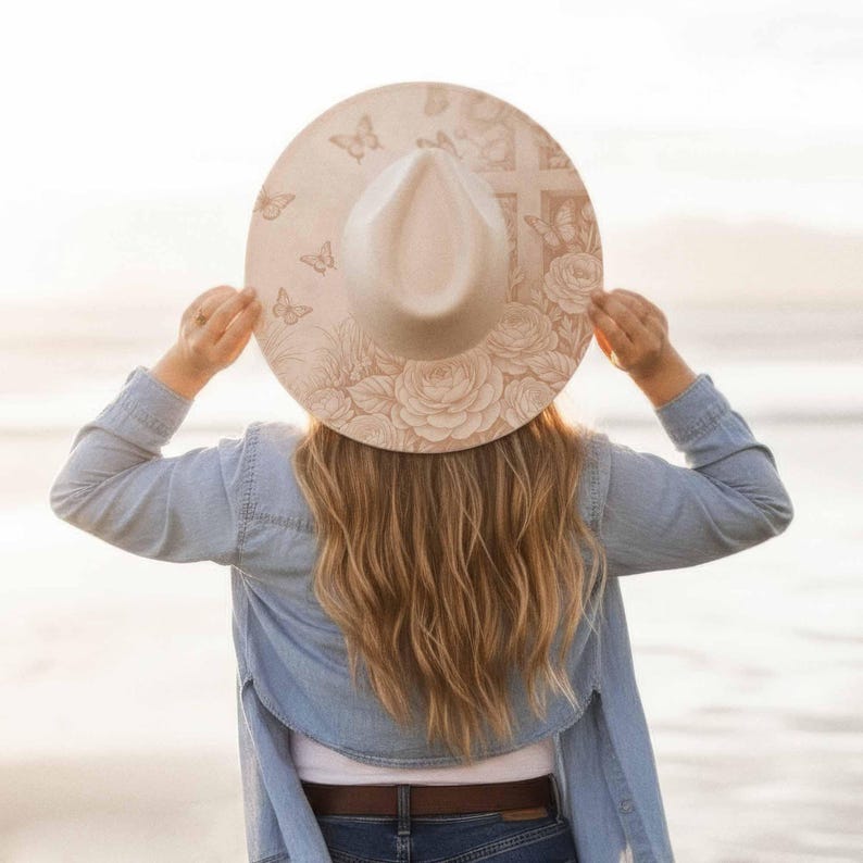 May include: A cream-colored hat with a wide brim, adorned with floral and butterfly designs. The hat features a cross design and roses. The hat is being held up by a person wearing a denim shirt.