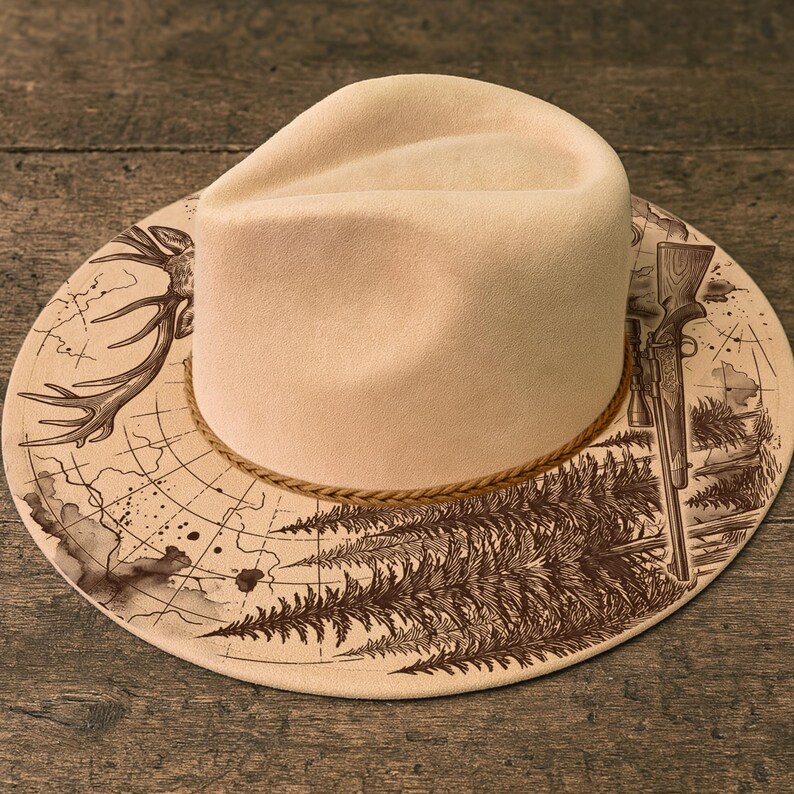 May include: A cream-coloured fedora-style hat with a wide brim featuring a detailed illustration. The brim displays a deer antler, a rifle, and a forest scene in brown ink. A braided rope encircles the base of the hat.