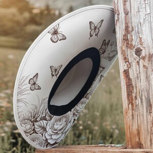 May include: A white sun hat with a black trim, adorned with brown butterfly and floral illustrations. The hat features a wide brim and is resting on a wooden surface, showcasing a nature-inspired design.