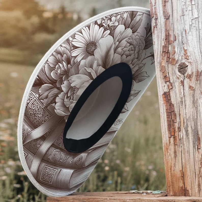 K&ouml;nnte beinhalten: Ein dekorativer Cowboyhut mit Blumen- und Buchdesign. Der Hut zeigt eine detaillierte Illustration von Pfingstrosen und G&auml;nsebl&uuml;mchen in Braun- und Wei&szlig;t&ouml;nen. Der Rand ist mit einem wei&szlig;en Rand versehen und die Innenseite ist schwarz.