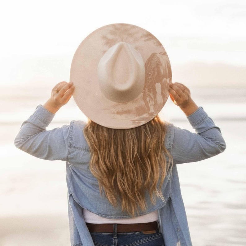K&ouml;nnte beinhalten: Eine Person h&auml;lt einen breitkrempigen, hellbeigen Hut mit cremefarbenem Oberteil hoch. Der Hut hat ein dezentes Design auf der Krempe. Die Person tr&auml;gt ein Jeanshemd und Jeans. Der Hintergrund ist eine verschwommene Strandszene.