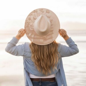 K&ouml;nnte beinhalten: Ein beiger Fedora-Hut mit breiter Krempe, verziert mit einem Blumen- und Buchdesign. Der Hut wird von einer Person mit langen blonden Haaren gehalten, die ein Jeanshemd tr&auml;gt. Das Design des Hutes umfasst einen Stapel B&uuml;cher und Blumen.