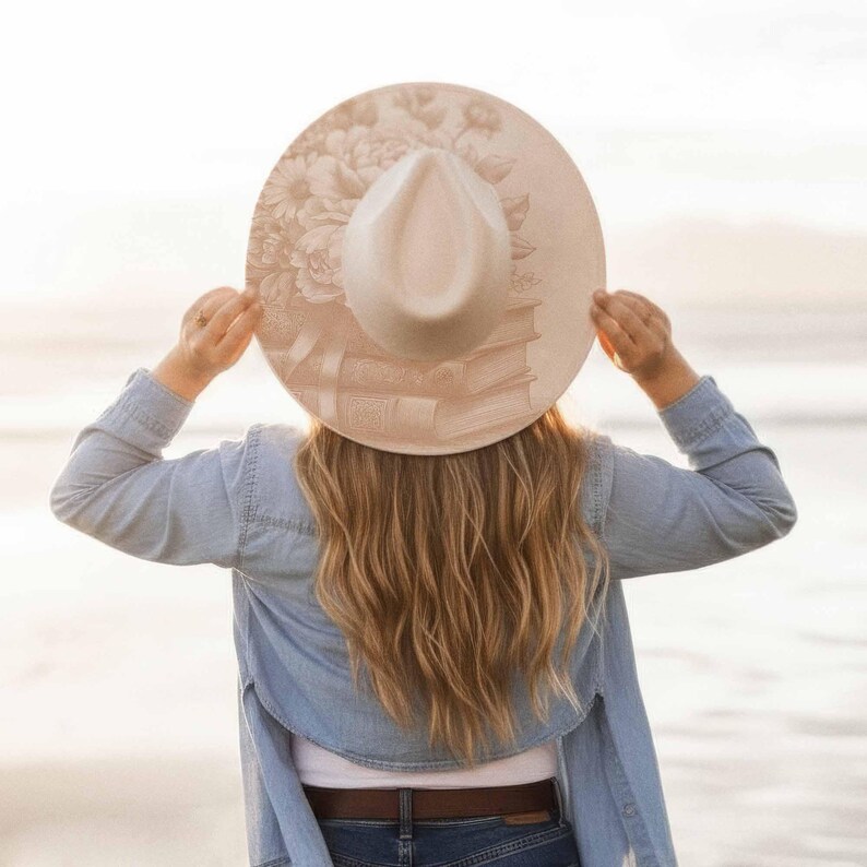 K&ouml;nnte beinhalten: Ein beiger Fedora-Hut mit breiter Krempe, verziert mit einem Blumen- und Buchdesign. Der Hut wird von einer Person mit langen blonden Haaren gehalten, die ein Jeanshemd tr&auml;gt. Das Design des Hutes umfasst einen Stapel B&uuml;cher und Blumen.