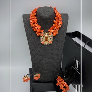 May include: A coral necklace, bracelet, and earrings set with a gold pendant featuring a large, clear gemstone. The jewelry is displayed on a black mannequin and a black box.