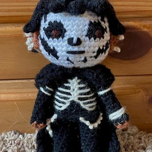 May include: A crocheted doll with a black and white skeleton costume. The doll has a white face with black eyes and a black mouth. The doll is wearing a black hooded cloak.