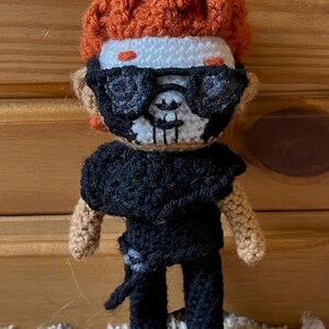 May include: A crocheted doll with orange hair, white face, black sunglasses, and a black skull mask. The doll is wearing a black shirt and black pants.