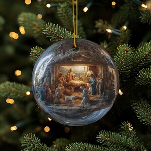 Puede incluir: Una bola de Navidad de cristal transparente con una cuerda dorada, que representa una escena de la Natividad. La obra de arte en el interior muestra figuras con túnicas reunidas alrededor de un bebé en un pesebre, con un cordero. La bola cuelga sobre un árbol de Navidad.