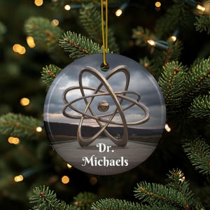以下が含まれることがあります： 風景を背景に、銀色の原子モデルが描かれた丸いクリスマスオーナメント。「Dr. Michaels」の文字が白で表示されています。オーナメントは金の紐から吊り下げられています。