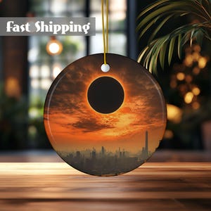 May include: A round ceramic ornament featuring a cityscape silhouetted against a dramatic sunset with a total solar eclipse. The sun is a bright orange and yellow, with a dark circle in the center.