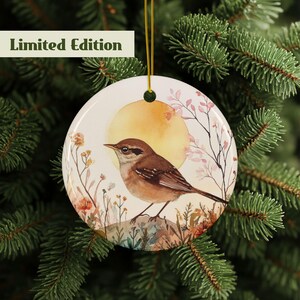 Könnte beinhalten: Runder Keramik-Weihnachtsschmuck mit einer Aquarellillustration eines Vogels auf einem Felsen, vor einem gelben Sonnenhintergrund. Der Schmuck hat eine goldene Aufhängeschnur und den Text "Limited Edition" in einem grünen Feld.
