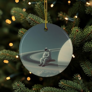 Op de afbeelding: Een ronde kerstversiering met een astronaut die op een planeet met ringen zit. De astronaut draagt een wit ruimtepak. De achtergrond toont een planeet met ringen en een donkerblauwe lucht. De versiering hangt aan een gouden draad.