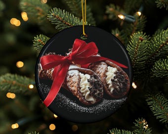 May include: A round, black Christmas ornament featuring a photograph of two cannoli pastries with white cream filling and chocolate chips, tied with a red ribbon. The ornament hangs from a gold string against a background of Christmas tree branches.